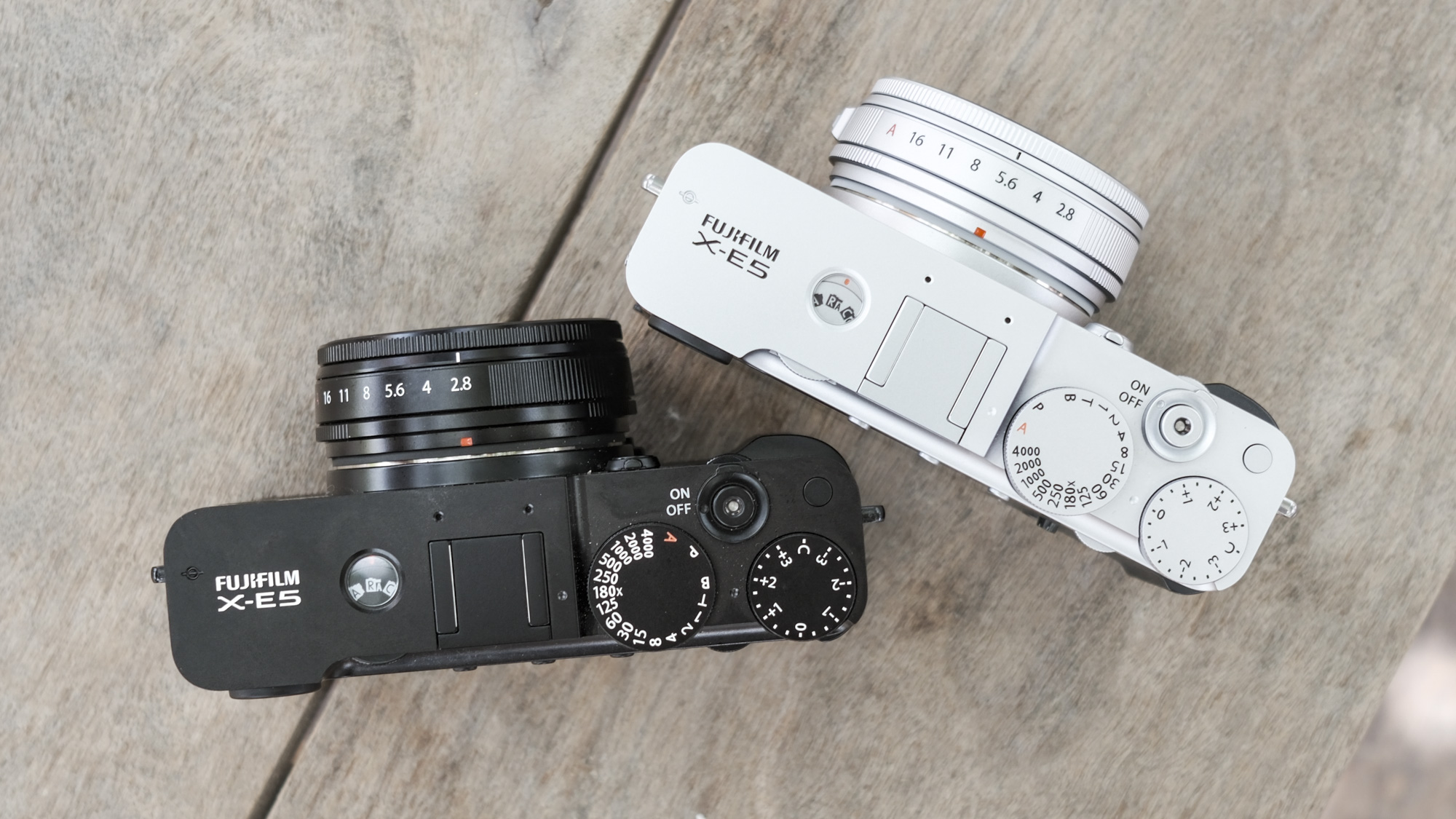 At the top of all black Fujifilm X-E5 alongside Silver Fujifilm X-E5, on a wooden bench