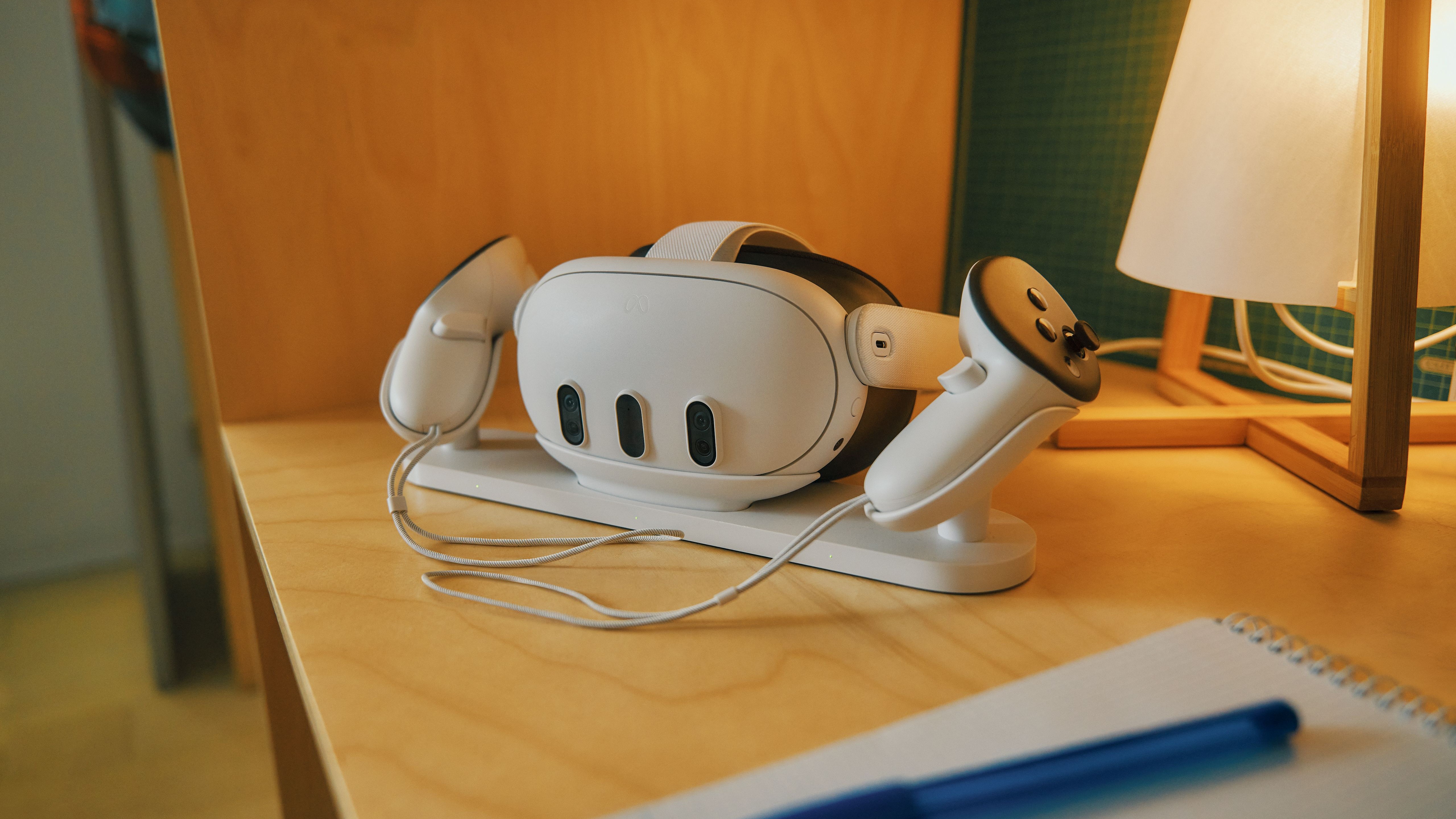 The Meta Quest 3 and controllers on their charging station, which sits on a wooden desk next to a lamp
