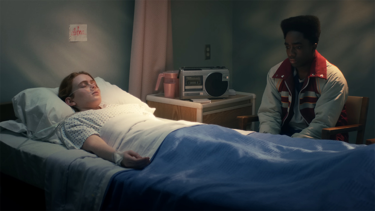 A comatose Max lying in a hospital bed while Lucas sits next to her in Stranger Things season 5
