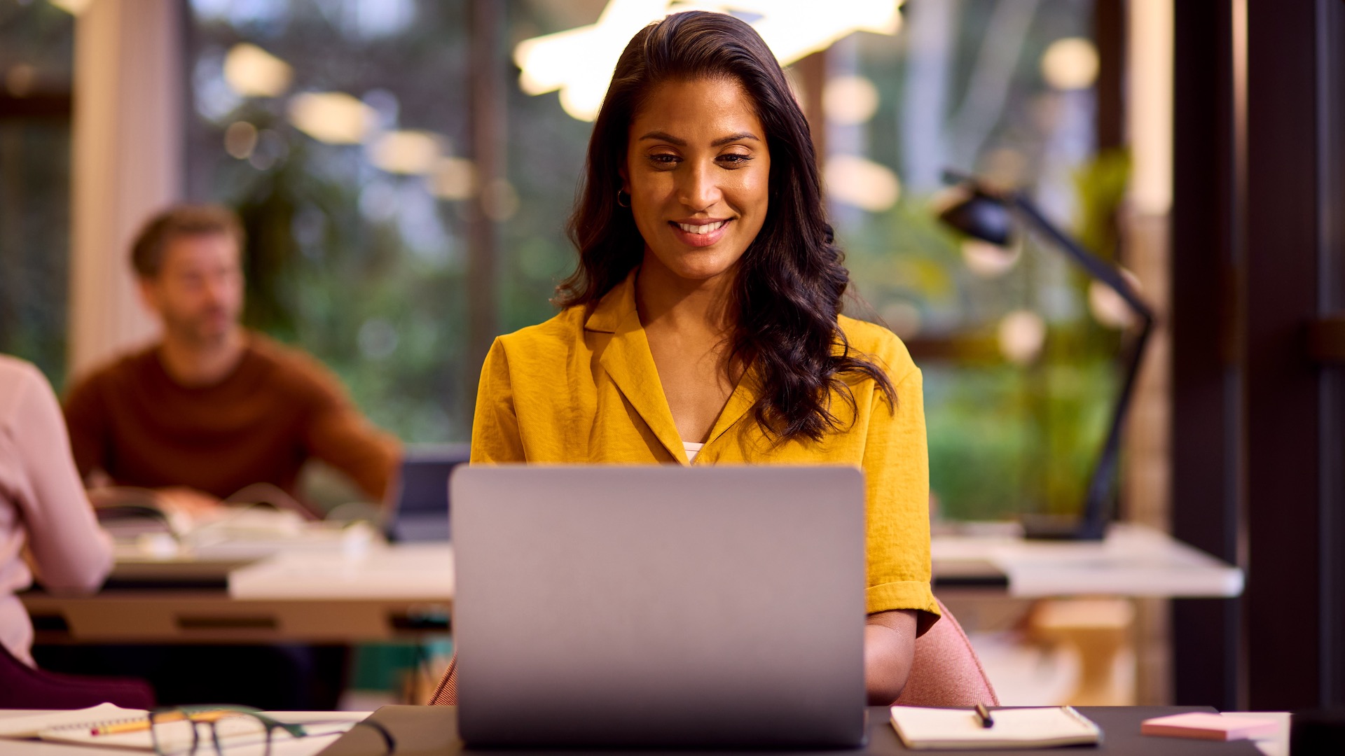 A young woman works on a Windows 11 laptop in a relaxed office.