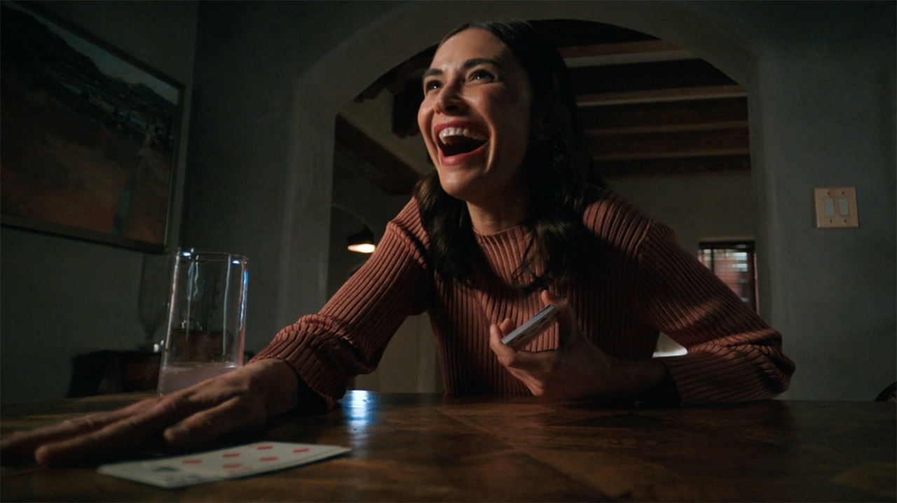 Zosia smiles widely and places her right hand on a pile of table cards in episode 8 of Pluribus