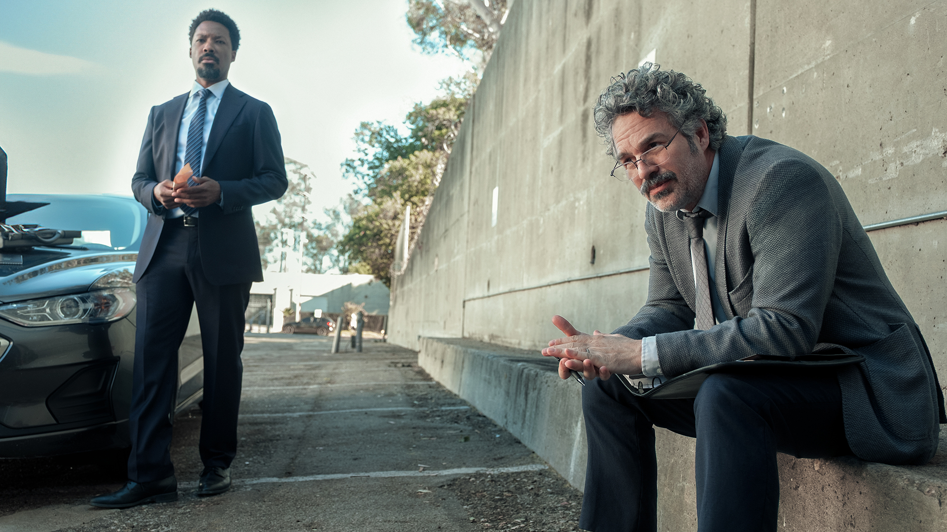 Detective Tillman standing next to a car while Detective Lubesnick sits on a nearby wall in Crime 101.