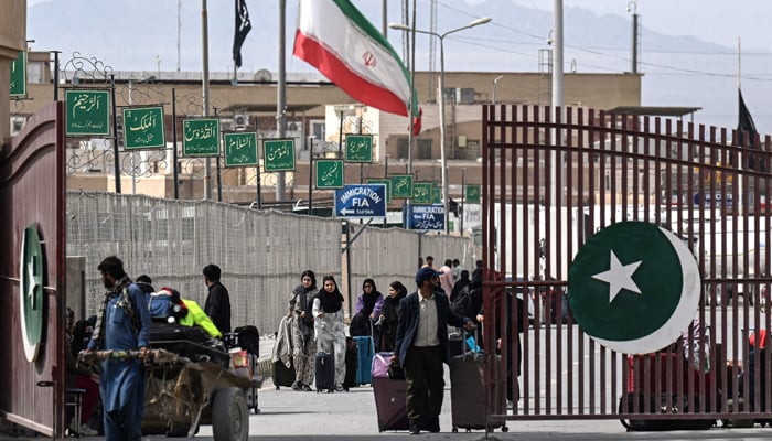 Pakistani nationals cross the Pakistan-Iran border after returning from Iran as an Iranian national flag (center) flies at half-mast in Taftan, Balochistan province, March 2, 2026, following the death of Iranian Supreme Leader Ayatollah Ali Khamenei amid US-Israeli strikes. -AFP