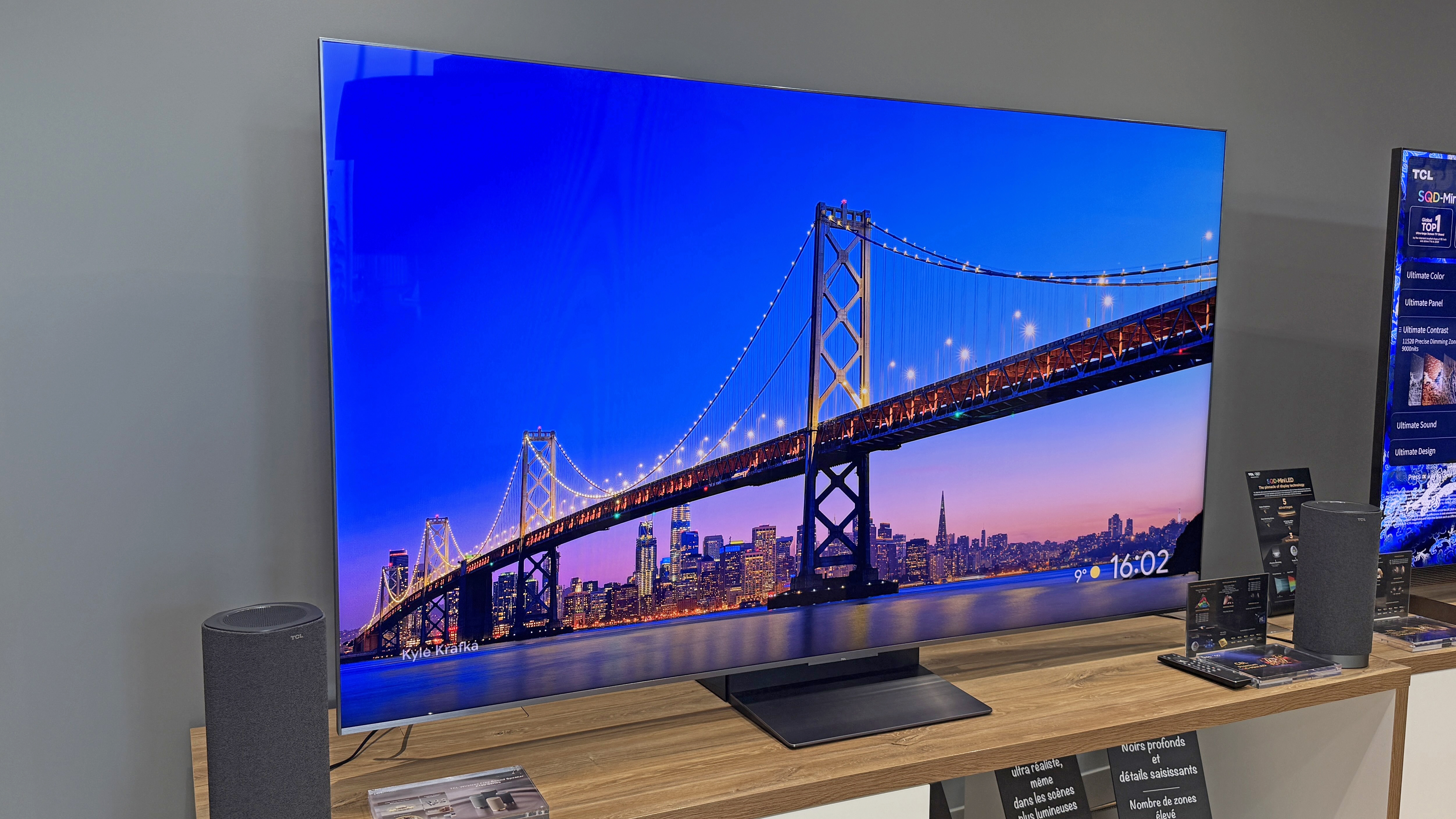The TCL C8L TV at a launch event, showing a striking blue image of a large and sky behind it