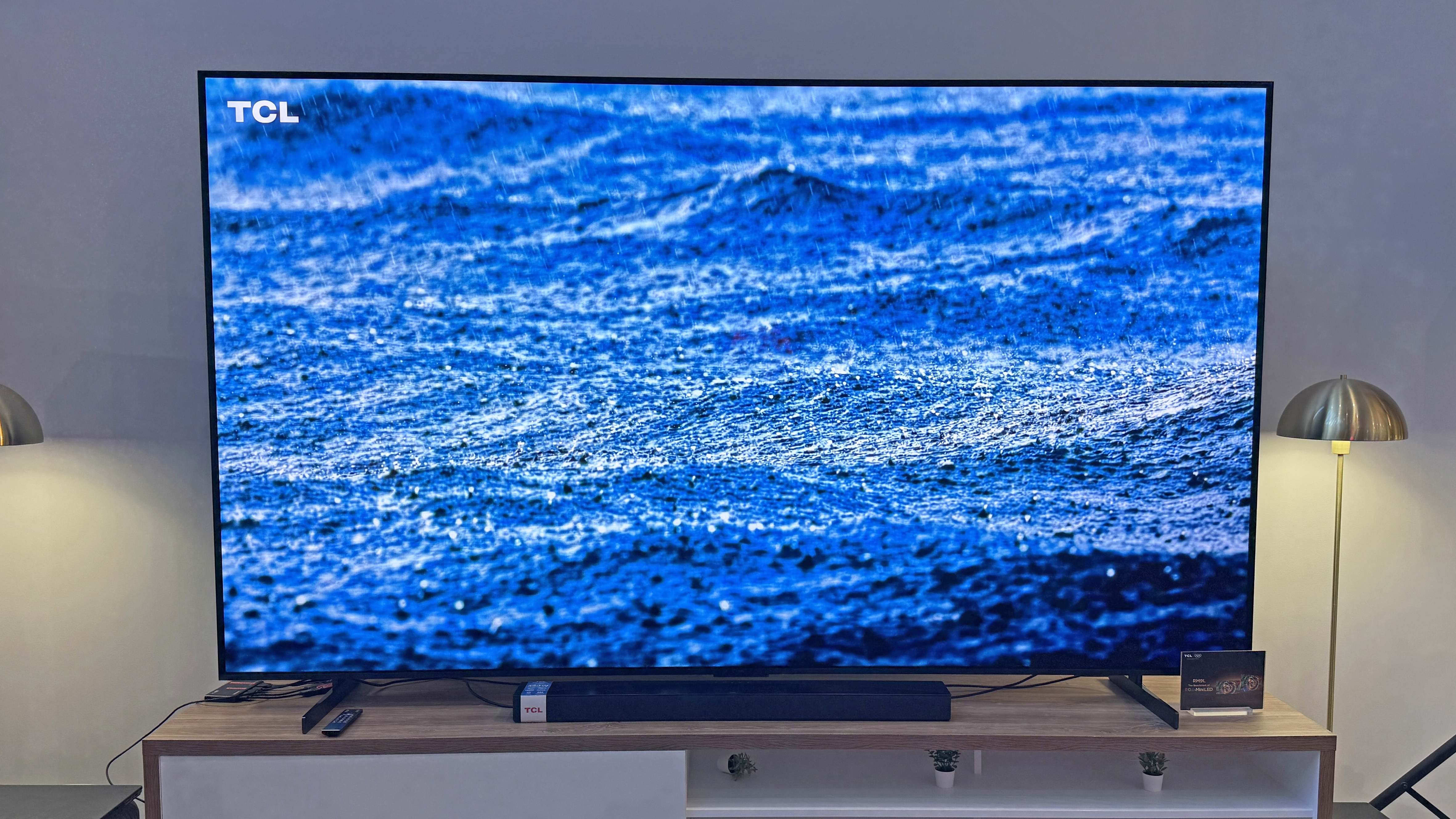 The TCL RM9L TV at a launch event, showing an image of rain falling on the ocean, with a striking blue tone and great detail in the rain splashes