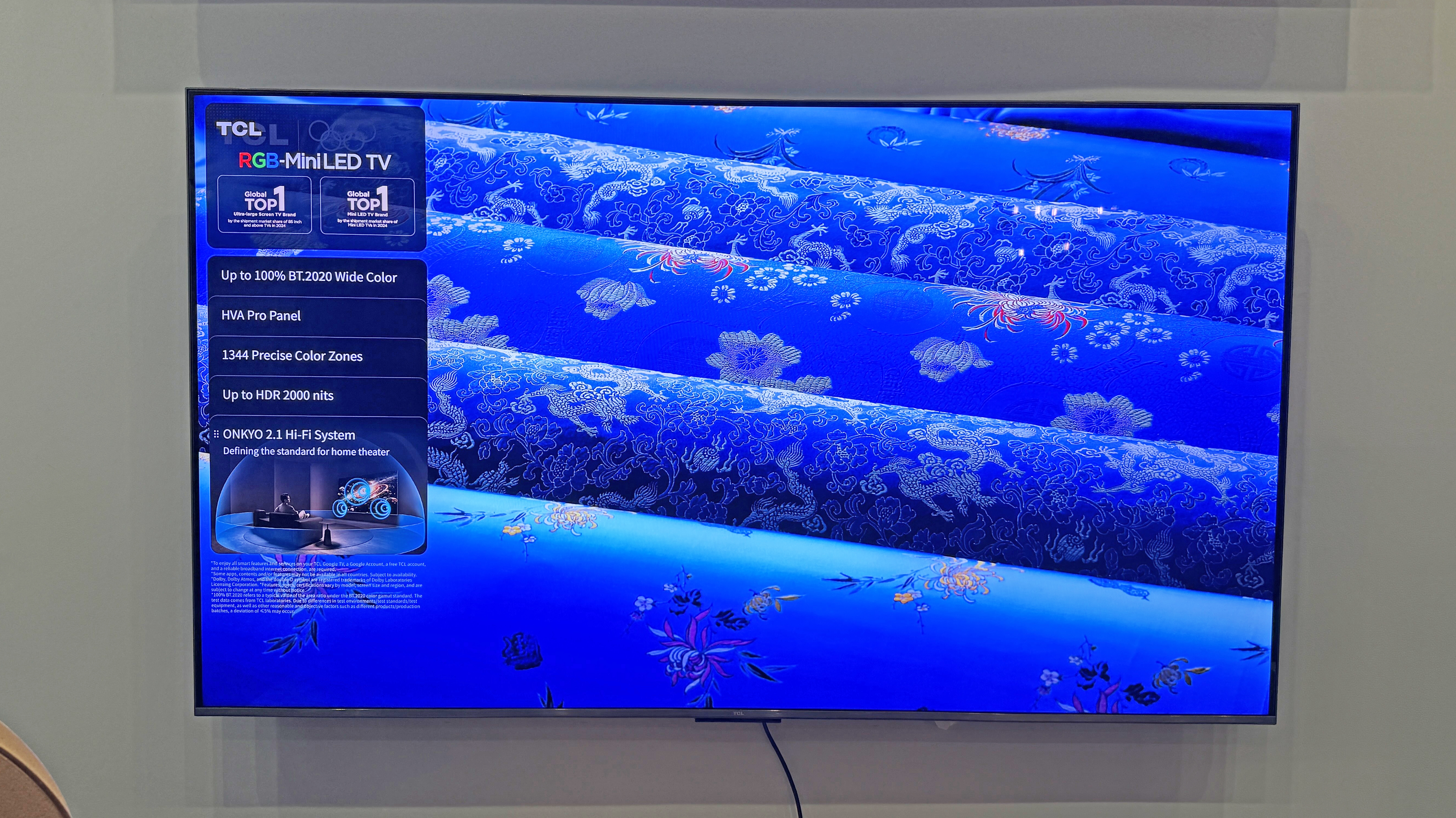 The TCL RM7L TV at a launch event, showing an image of rolls of blue fabric, with a bright, electric tone for the blue color