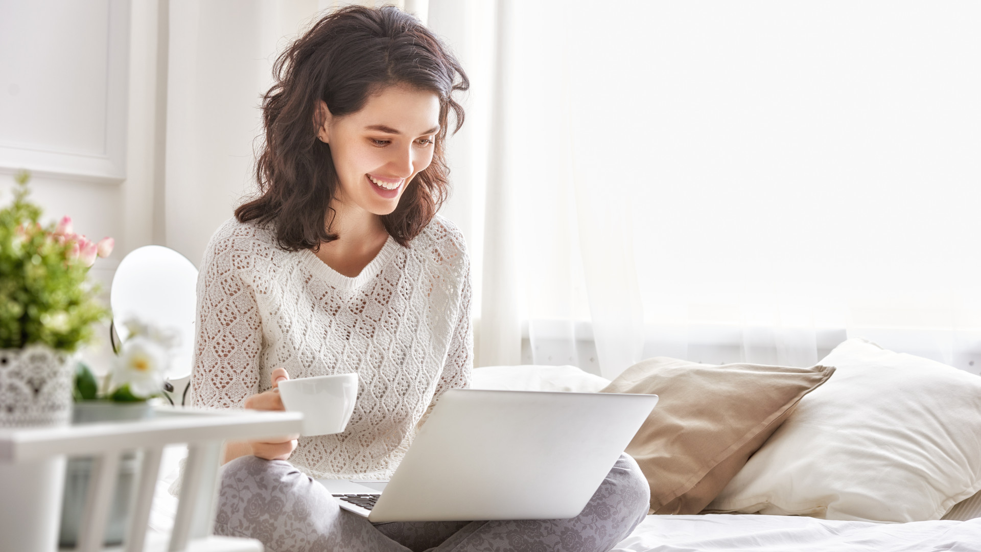Happy woman sitting on bed with coffee and Windows 11 laptop