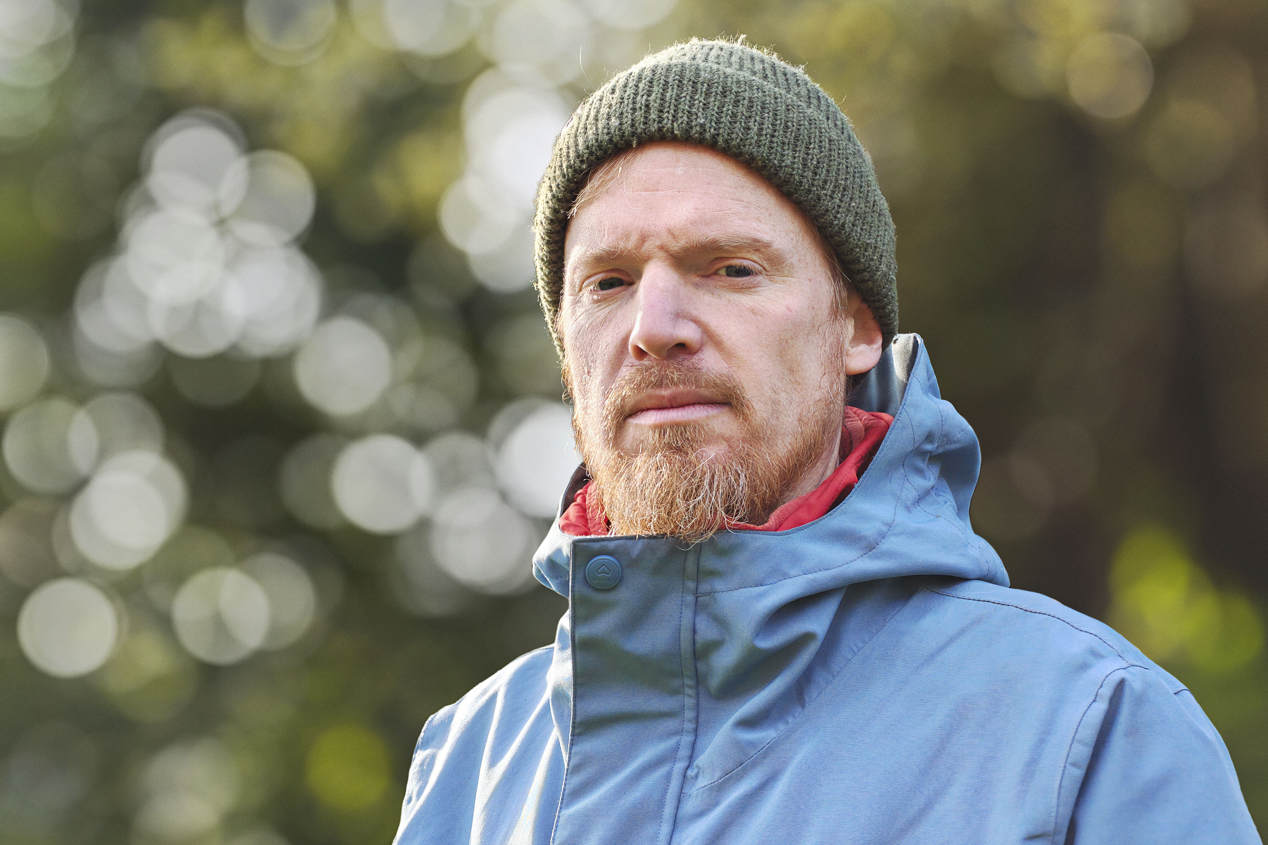 Portrait of a man in blue coat and green cap, with dappled light through a tree in the background