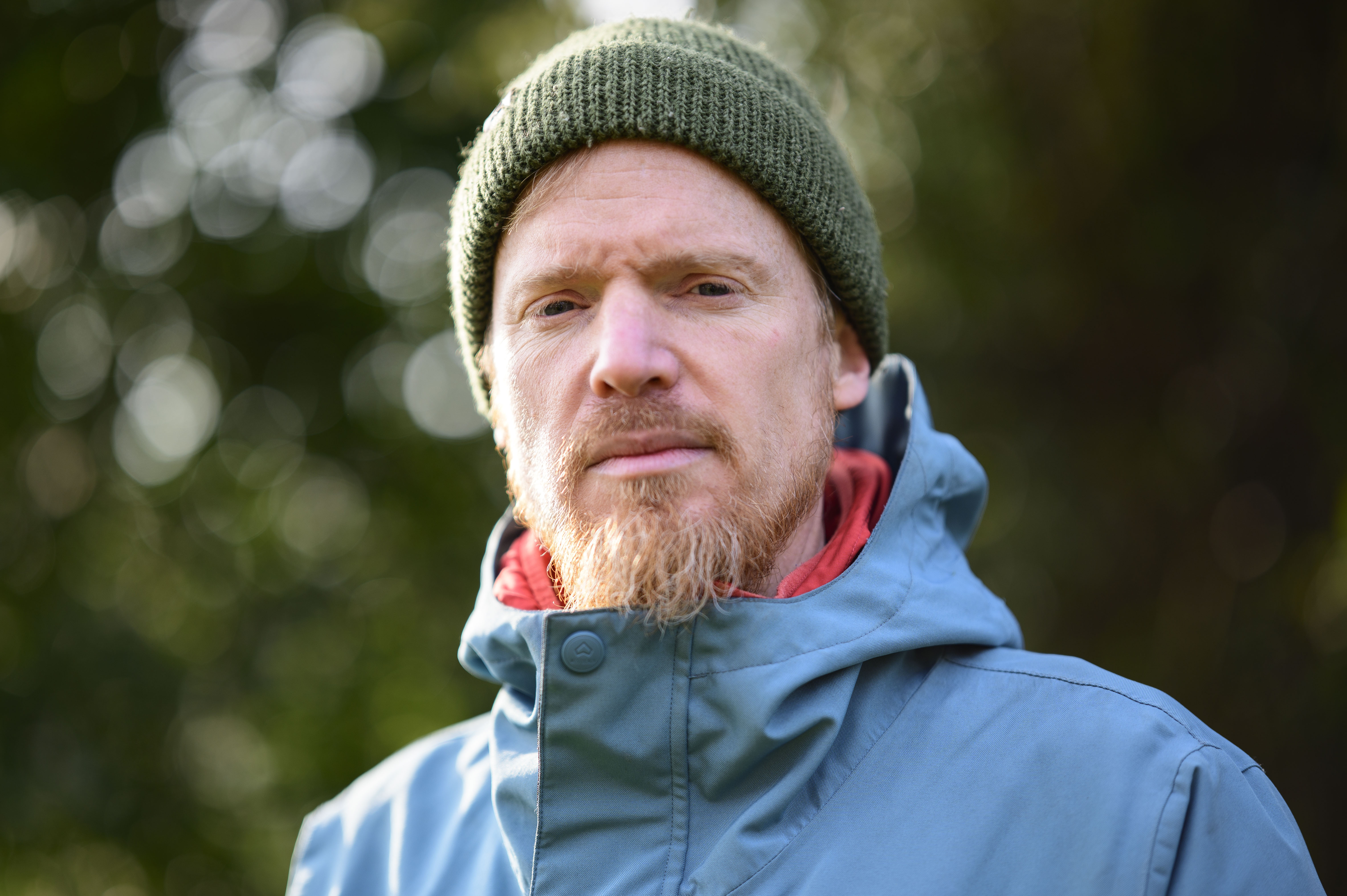 Portrait of a man in blue coat and green cap, with dappled light through a tree in the background