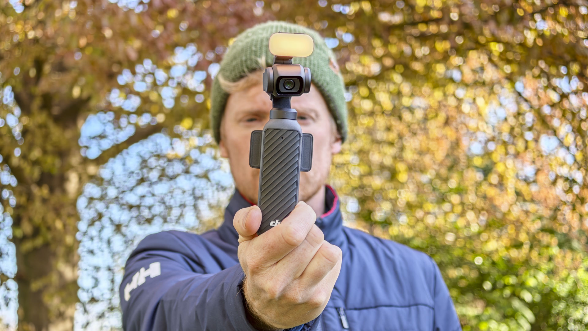 Man in beanie holding DJI Osmo Pocket 4 vlogging camera with its magnetic fill light, an autumnal tree behind him