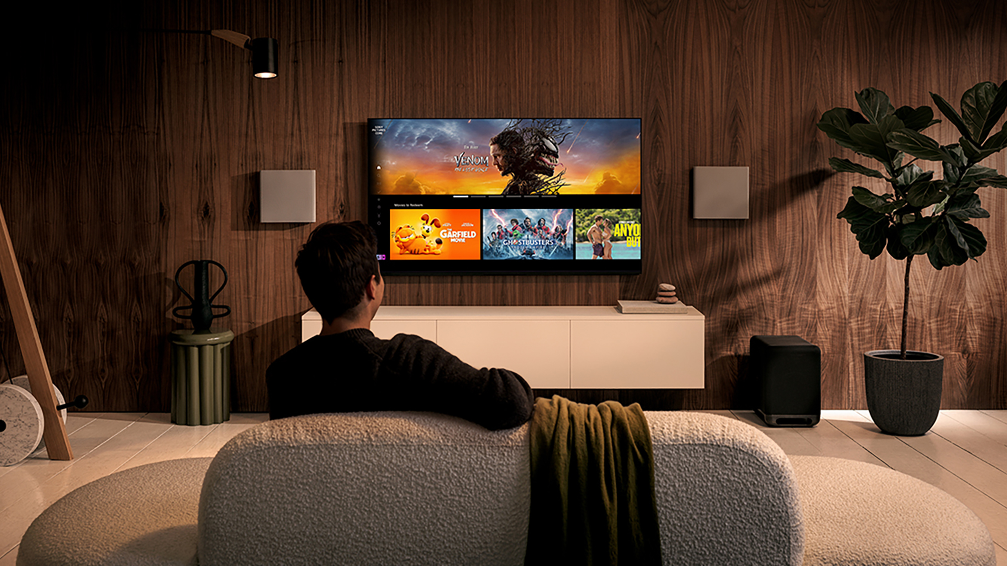 A man seen from behind, sitting on a sofa, watching a television displaying thumbnails of selected content.