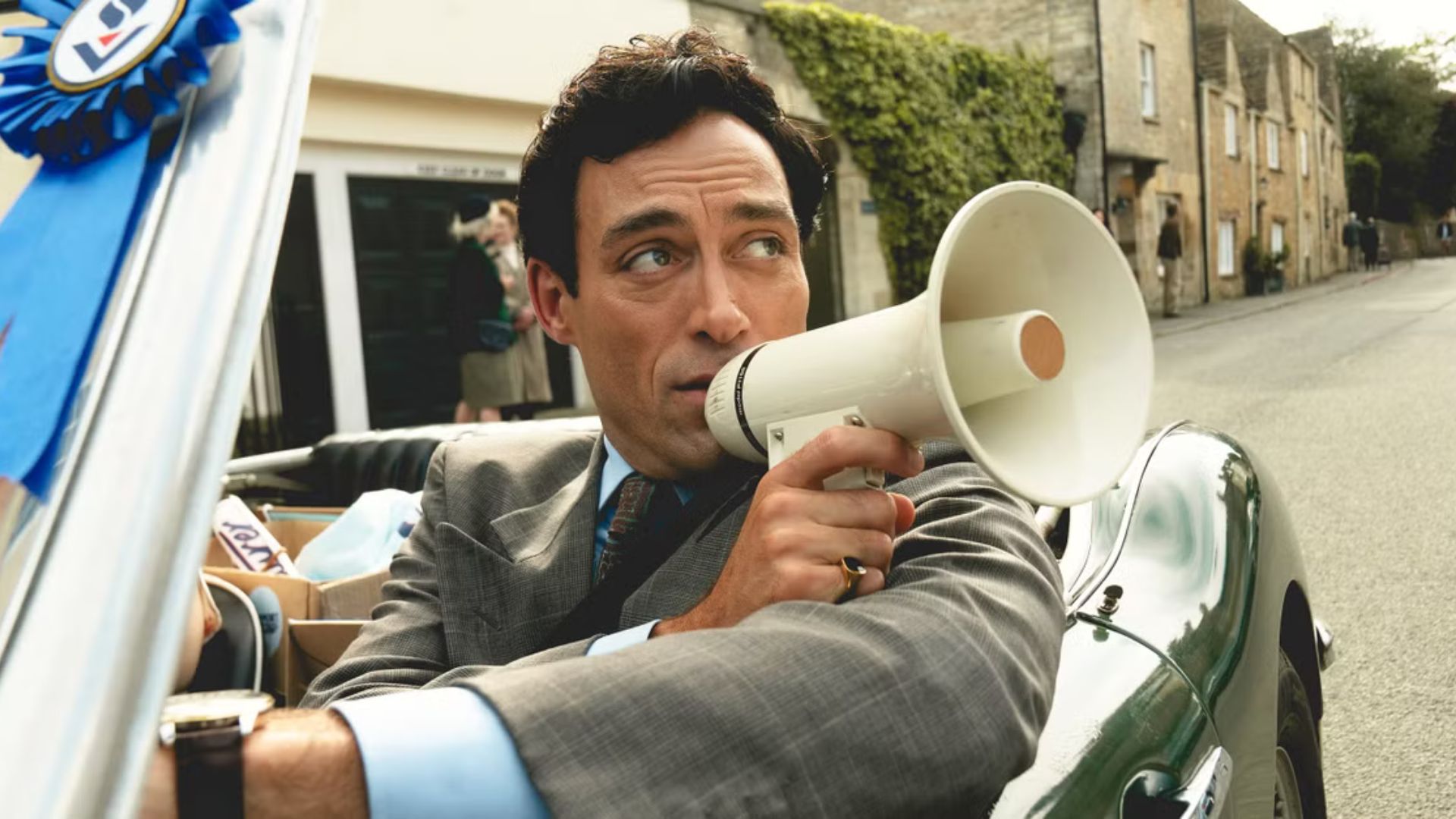 Rupert Campbell-Black speaks into a megaphone while driving a convertible