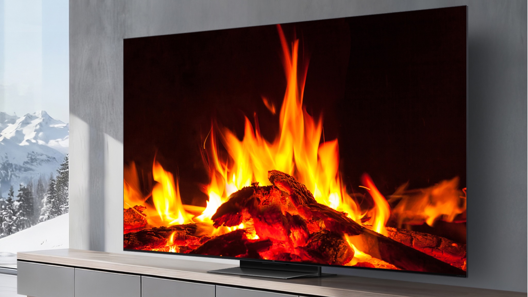 A TCL C7L TV showing a wood fire. It's in a gray decorated room with snow-capped mountains visible through a floor-to-ceiling window to the left of the TV stand.