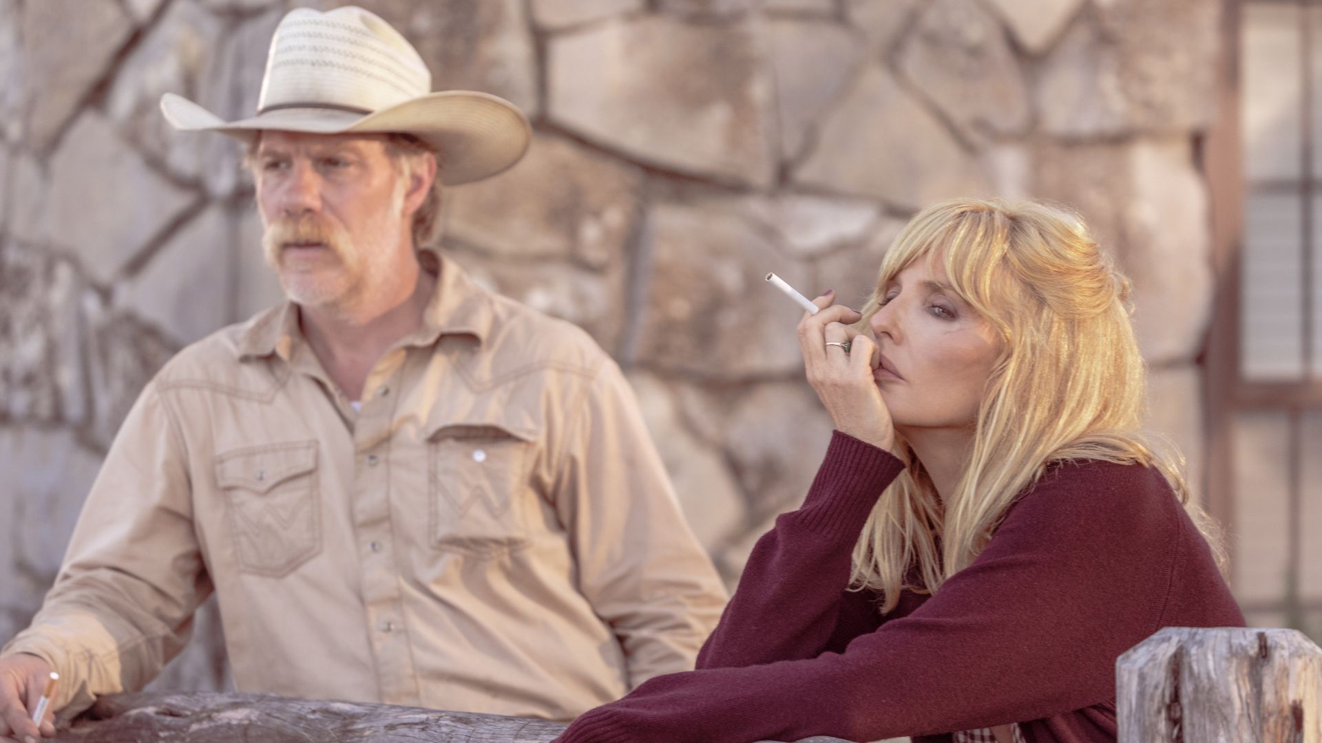 Beth smokes while leaning against a wooden fence with Zachariah standing behind.