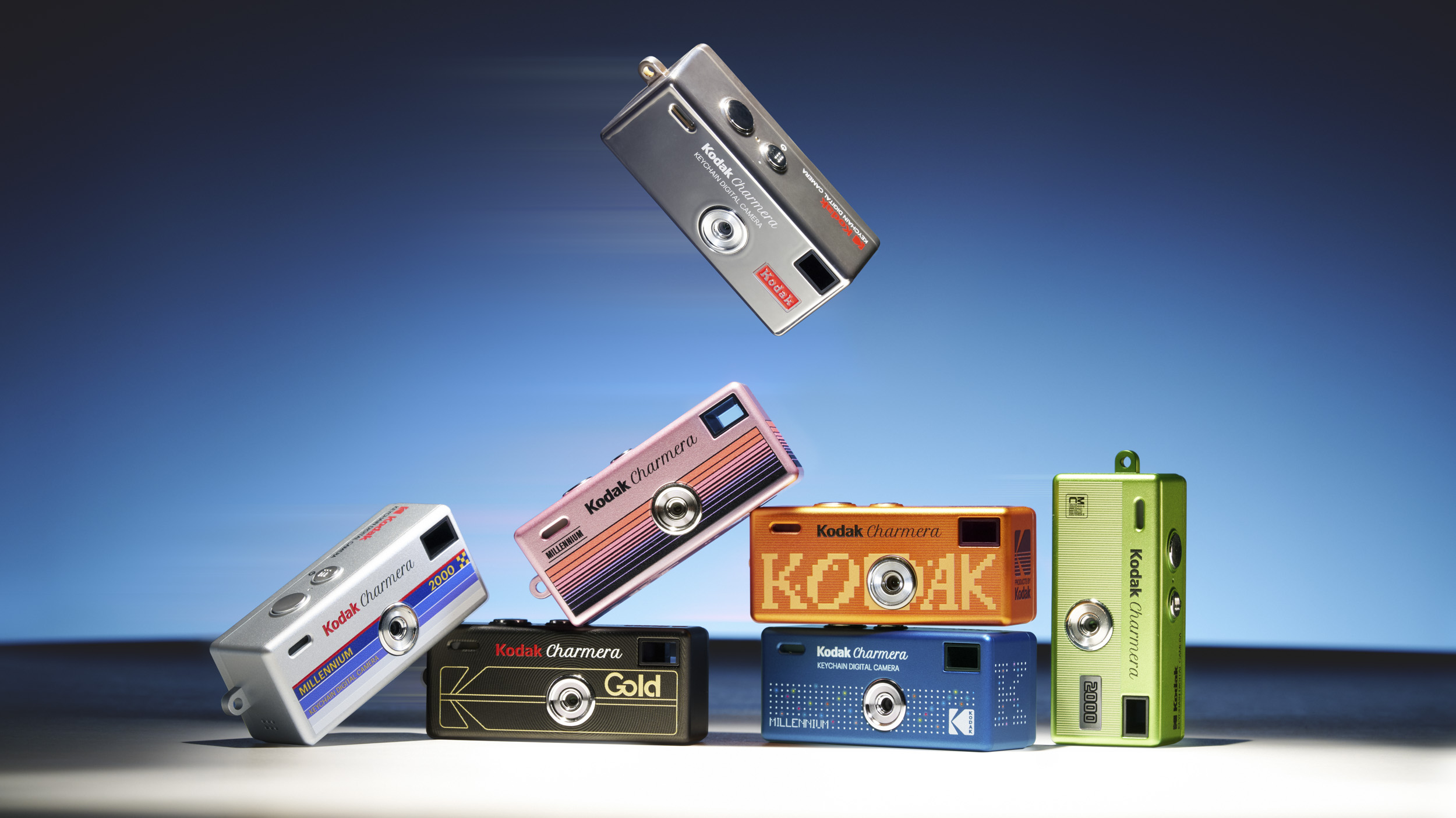 Seven different Kodak Charmera Millennium Edition cameras in a studio on a white surface with a bright blue background