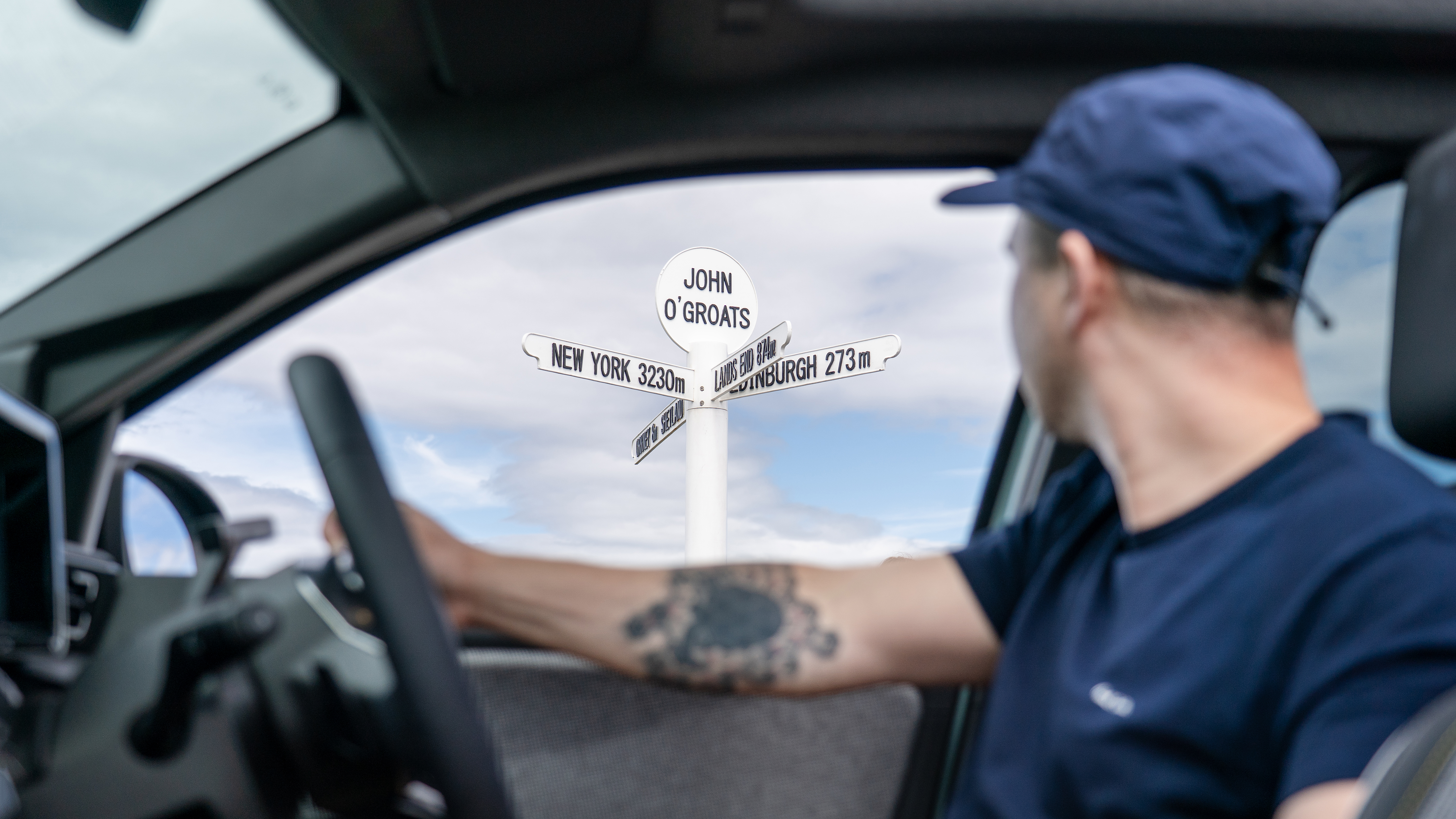 A man looking out of the window of an electric vehicle at a sign for John o'Groats