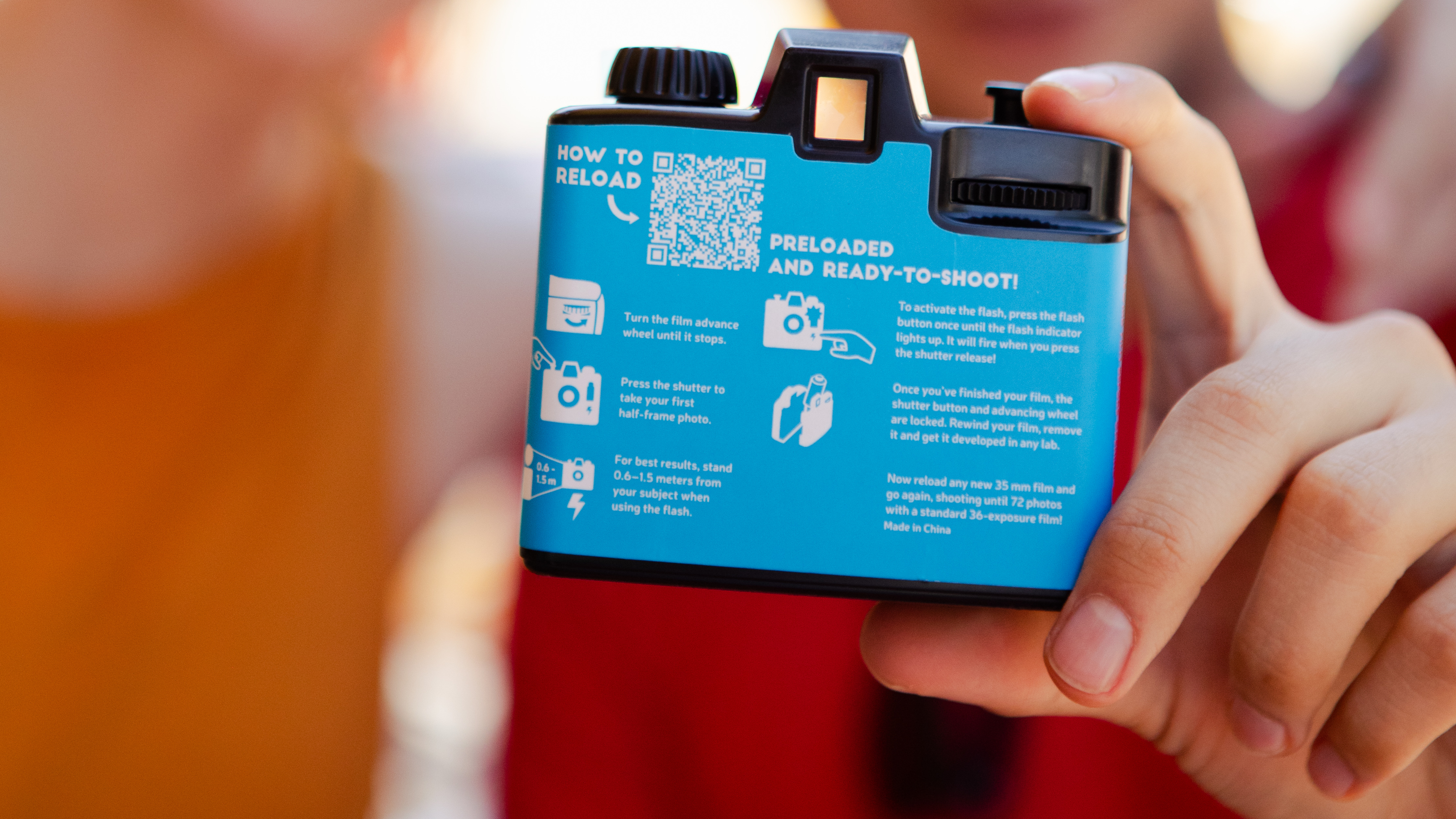 Photograph of a hand holding the Lomography single-use rechargeable half-frame film camera, showing the back of the camera with written instructions, diagrams and a QR code